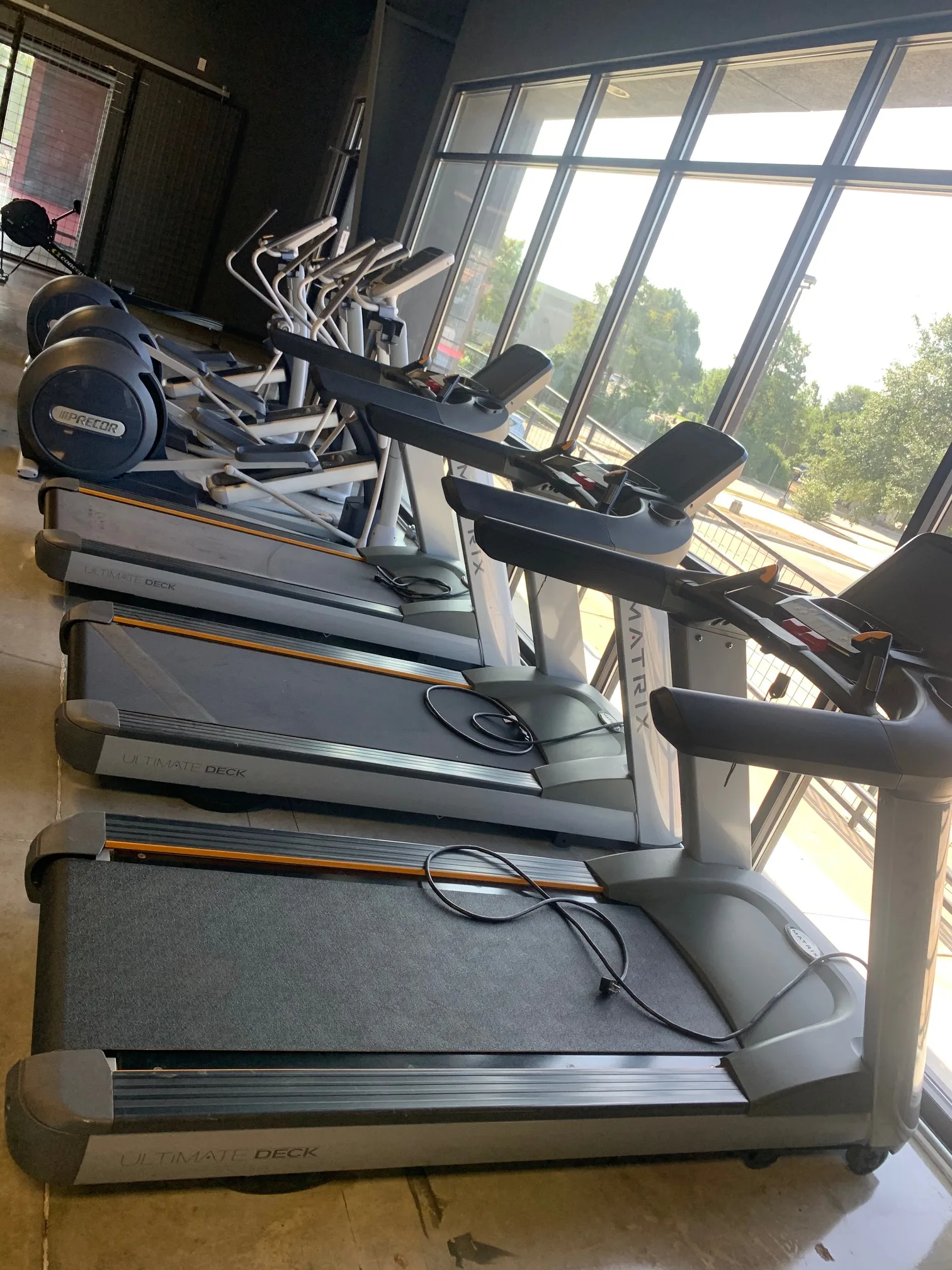 Three Matrix treadmills in a row along the floor-to-ceiling front glass, Precor ellipticals behind them